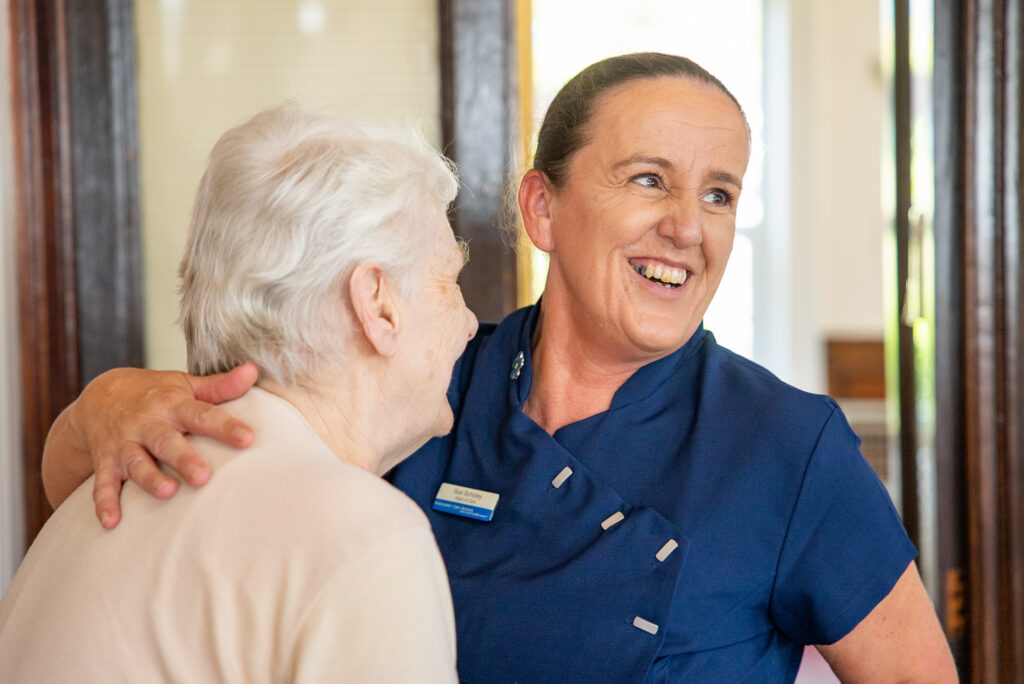 A close-up, heartwarming portrait at Cartmel Grange care home showing Head of Care Sue Scholey in a dark blue tunic uniform. She smiles broadly with joy while holding an elderly female resident in a warm embrace, with the resident's arm wrapped gently around her shoulder.