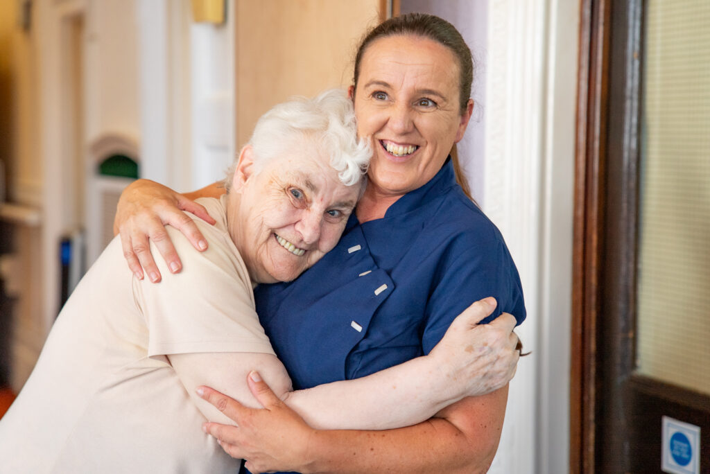 A heartwarming, candid moment in a carpeted hallway at Cartmel Grange care home. A female carer in a navy tunic and a senior female resident wearing a light beige t-shirt are embraced in a tight, affectionate hug, both smiling with happiness and comfort.