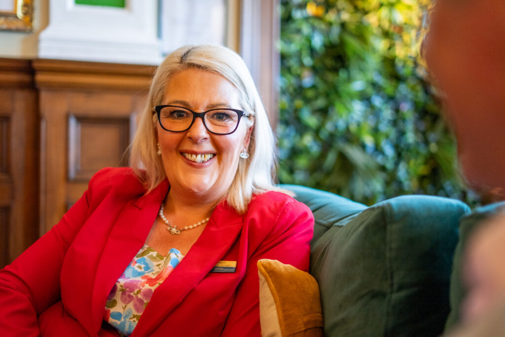 A warm indoor portrait of Registered Manager Shelly Ralph at Cartmel Grange care home. She has straight blonde hair, wears black-rimmed eyeglasses, a pearl necklace, a floral patterned top, and a vibrant red blazer with her staff name badge. She sits comfortably on a plush green armchair, smiling broadly during a conversation.