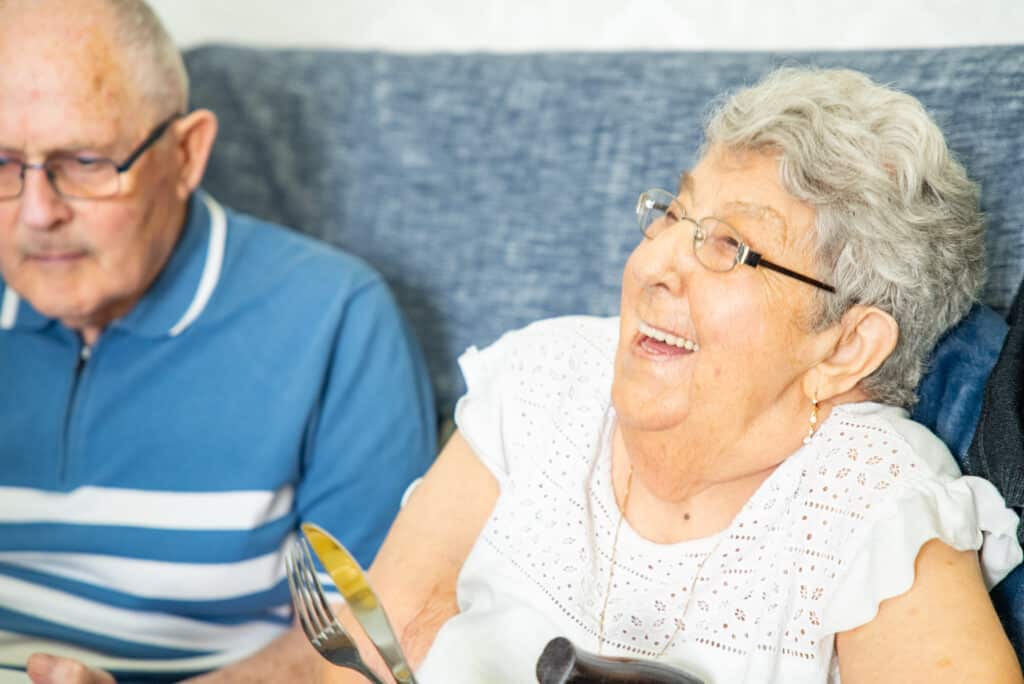 A warm, candid shot of an elderly female resident with short grey hair and glasses smiling brightly while holding a knife and fork at Clarendon Grange care home. She wears a white eyelet-patterned blouse and sits indoors, with an elderly man in a blue polo shirt sitting alongside her on a blue couch in the background.