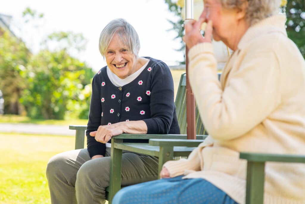 A candid outdoor shot at East Croft Grange care home showing an elderly woman with short grey hair laughing joyfully. She wears a black cardigan embroidered with small white and pink flowers, a white top, and grey trousers while sitting on a green wooden bench in a sunlit garden setting. In the foreground, another senior resident in a cream cardigan sits nearby, slightly blurred.