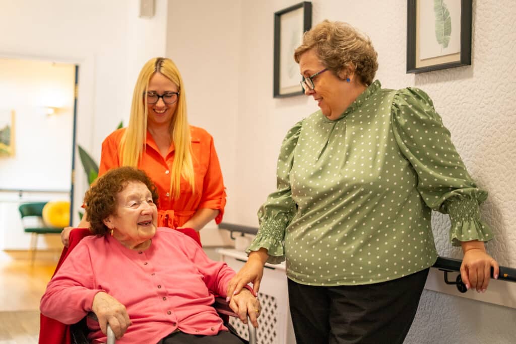A warm, cheerful interaction inside a hallway at East Croft Grange care home. An elderly woman in a wheelchair, wearing a pink cardigan, laughs joyfully as a female staff member in a green polka-dot blouse gently holds her hand and touches her shoulder in comfort. Behind the resident stands another staff member with long blonde hair and glasses, wearing a bright orange dress and smiling warmly.