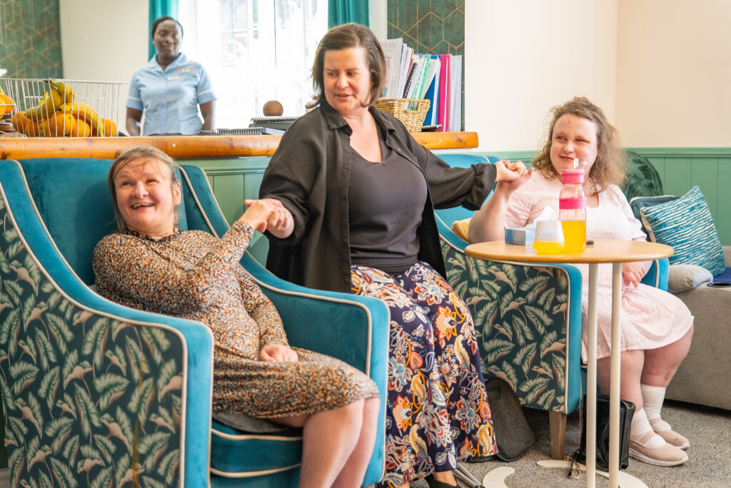 Residents and staff gather together in a brightly decorated lounge, holding hands and chatting warmly in plush teal patterned armchairs, illustrating the friendly, community-focused social environment provided at Linford Grange Care Home.